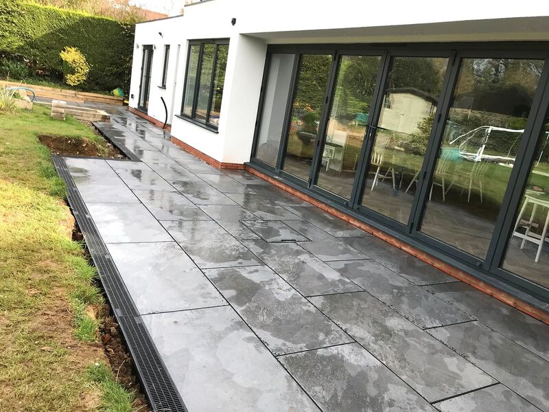 Grey porcelain paved terrace running along bi-fold doors with a drainage channel beside the lawn